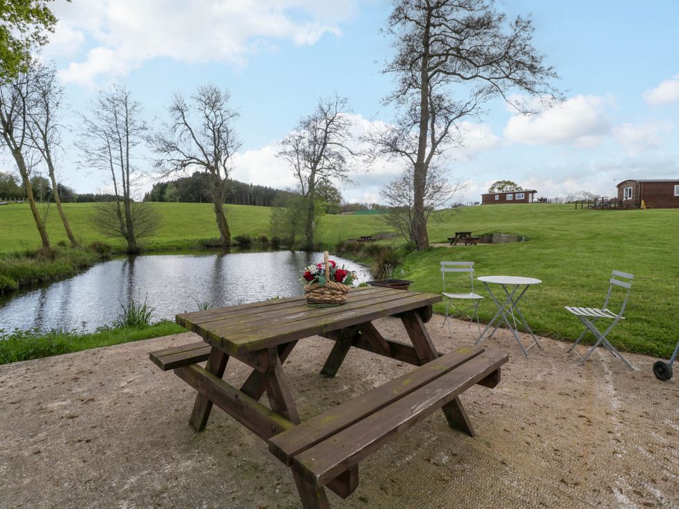 A wooden picnic table with a basket of flowers near a pond and green grassy field at Hollie Raglan