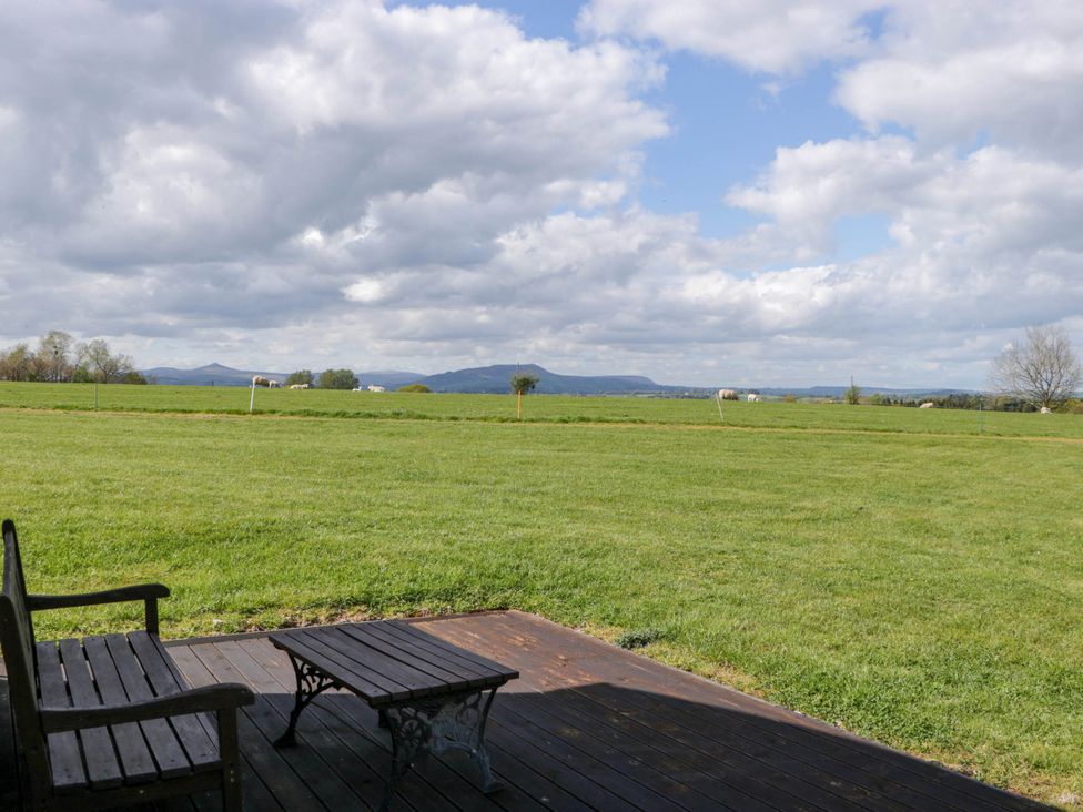 A wooden bench and table on a deck overlooking a grassy field with distant hills and clouds at Hollie in Raglan