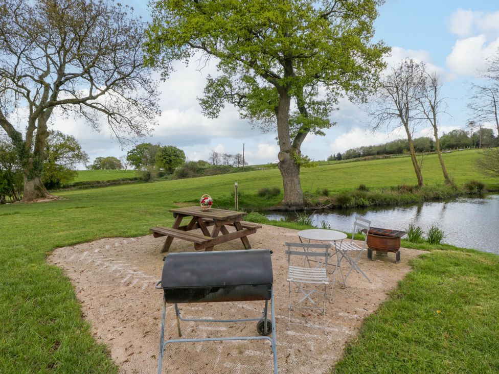 An outdoor seating area with a picnic table a round metal table two chairs and a barbecue near a pond in a grassy field at Hollie in Raglan