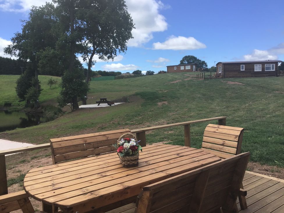 A wooden outdoor table with a flower basket on it surrounded by chairs on a deck overlooking a grassy hill with two wooden cabins and a picnic table near trees