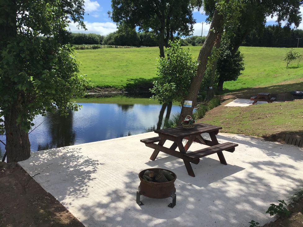 An outdoor area with a wooden picnic table and a fire pit near a small pond with grassy fields and trees in the background at Hollie in Raglan