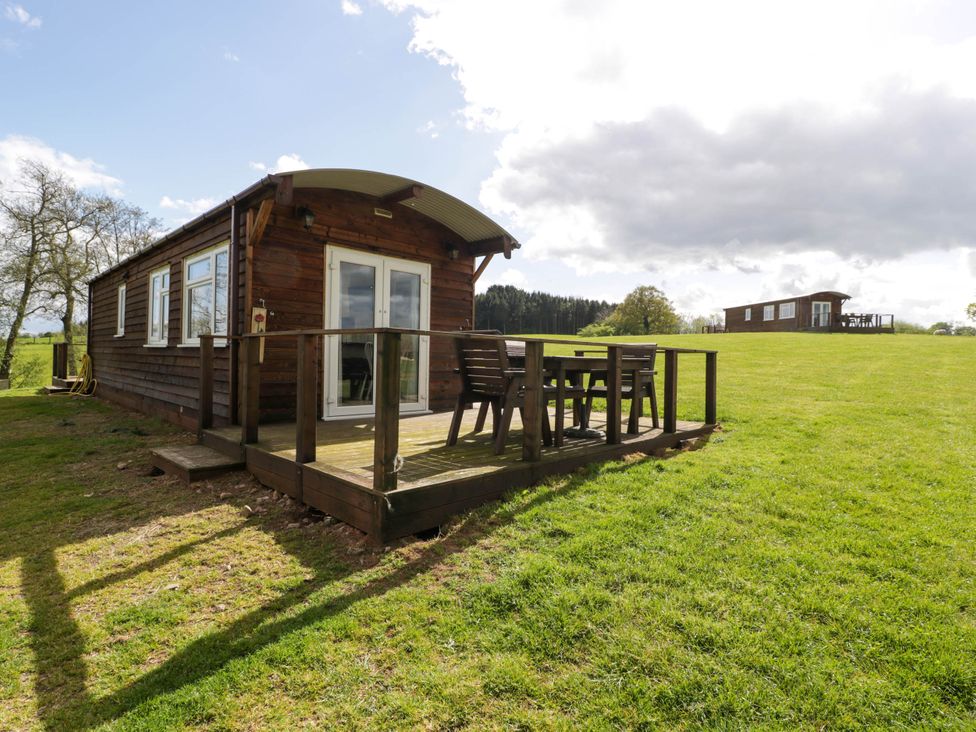 A wooden cabin with a deck and outdoor furniture on a grassy hill with another cabin in the distance at Poppy in Raglan