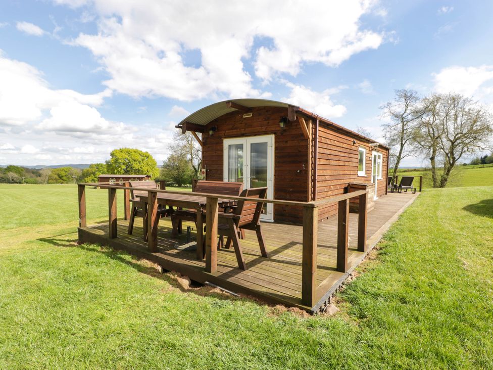 A wooden cabin with an outdoor dining table and chairs on a wooden deck in a grassy field at Poppy in Raglan