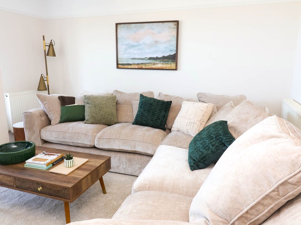 A living room with a beige sectional sofa with green and beige cushions a wooden coffee table with books and a plant and a floor lamp at Combe View in Ilfracombe