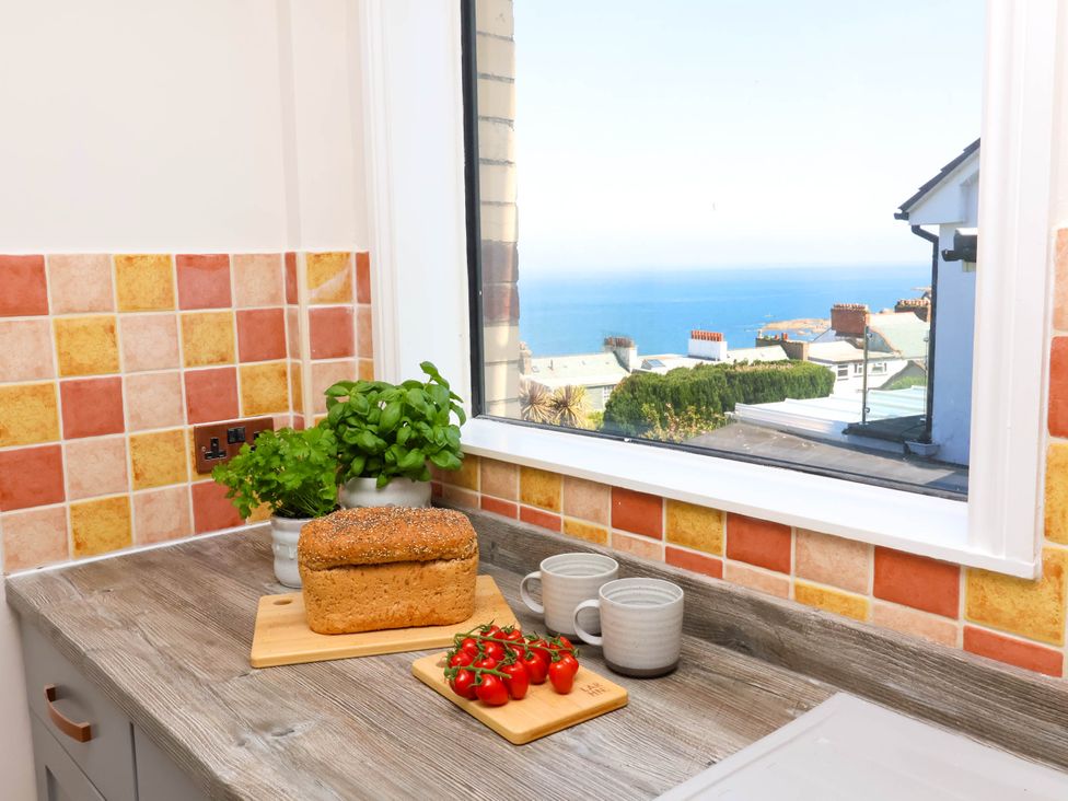 A kitchen countertop with a loaf of bread, two mugs, cherry tomatoes on a wooden board, potted herbs and a window showing a view of rooftops and the sea at Combe View in Ilfracombe