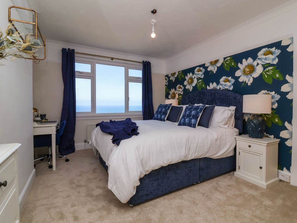 A bedroom with a double bed featuring white bedding and blue cushions against a floral wallpaper and a window overlooking the sea at Combe View in Ilfracombe