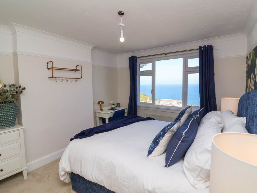 A bedroom with a bed pillows a window with ocean view a desk with lamp and a vase with flowers at Combe View in Ilfracombe