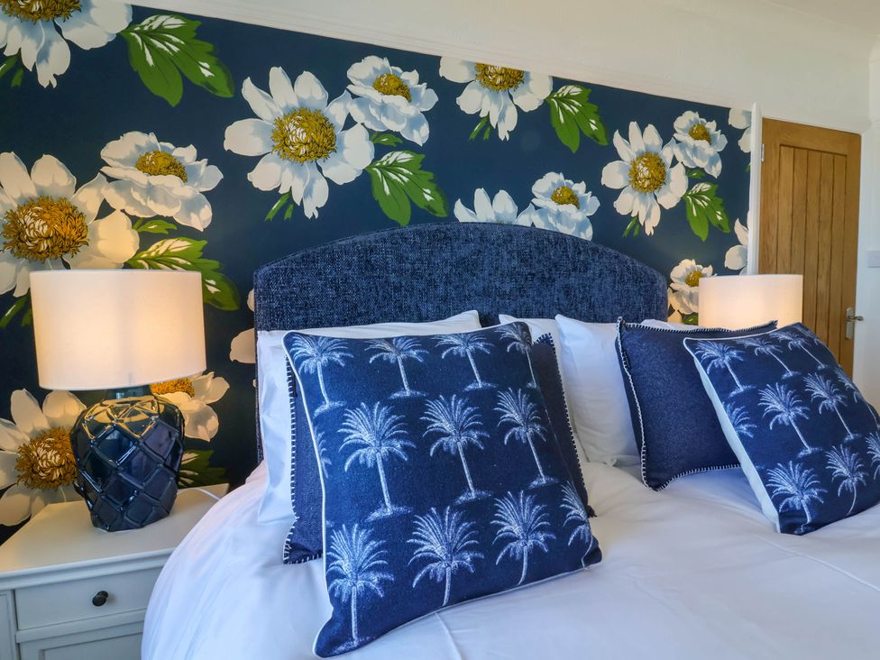 A bedroom with a bed featuring blue cushions with palm tree prints two bedside tables with lamps and a floral wallpaper at Combe View in Ilfracombe