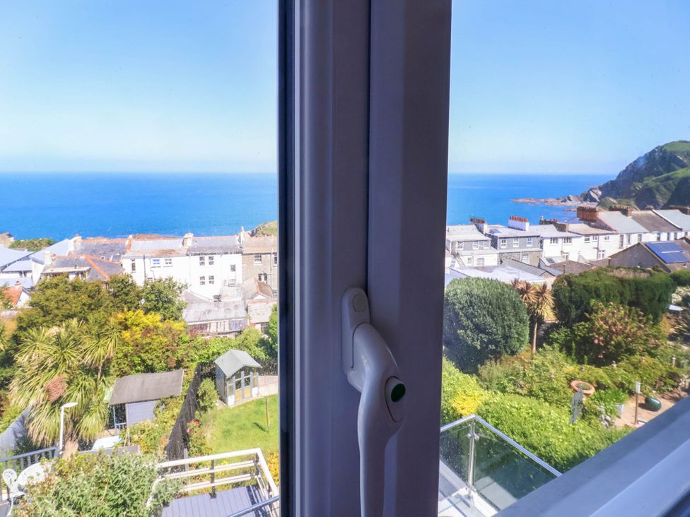 View through a window showing rooftops, gardens, and the sea with hills in the background at Combe View in Ilfracombe