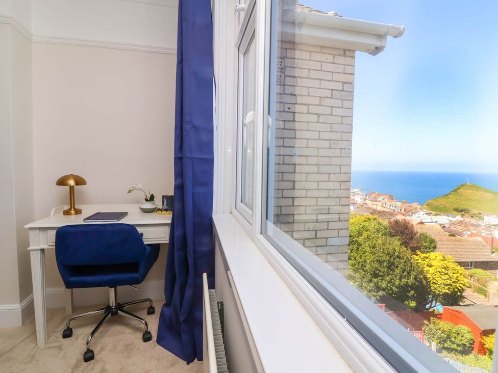 A desk with a lamp chair and notebook next to a window overlooking houses trees a hill and the sea at Combe View in Ilfracombe