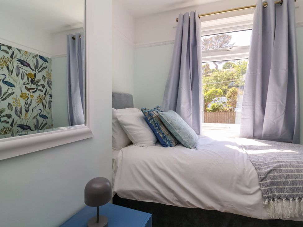 A bedroom with a bed with pillows and blanket near a window with curtains and a blue bedside table with a lamp at Combe View in Ilfracombe