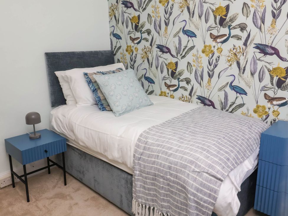 A bedroom with a single bed with pillows and a blanket next to blue bedside tables and a wallpaper with bird and leaf patterns at Combe View in Ilfracombe
