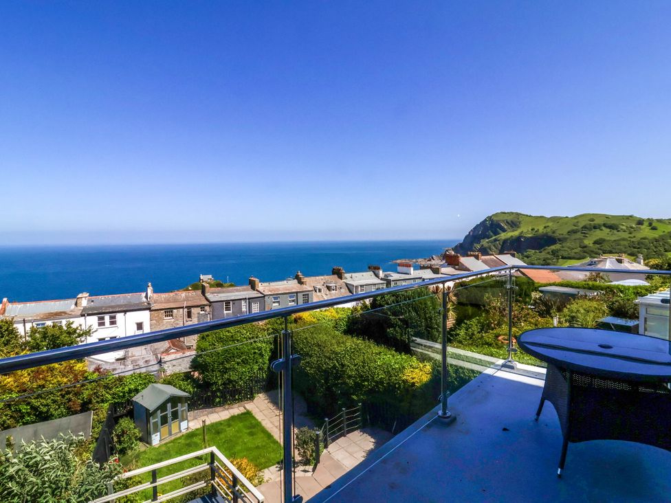 A balcony with a table overlooking houses gardens the sea and a green hill at Combe View in Ilfracombe