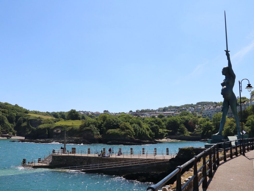 A coastal pier with people walking and fishing a tall statue holding a sword near a railing and houses on a hill in the background at Combe View in Ilfracombe