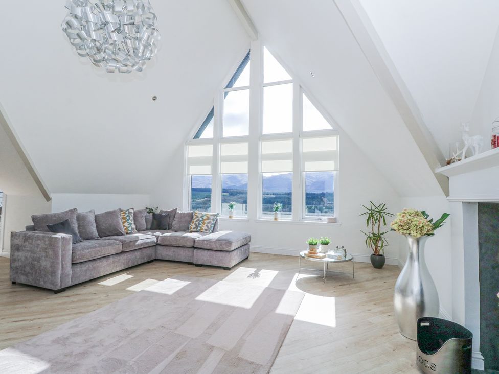 A living room with a grey sectional sofa large triangular windows and a metal vase with flowers at Macgregor Ranch in Achnabobane near Spean Bridge