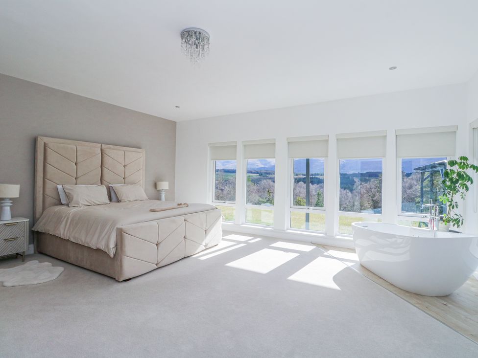A bedroom with a large bed nightstands a bathtub and multiple windows with a view at Macgregor Ranch in Achnabobane near Spean Bridge