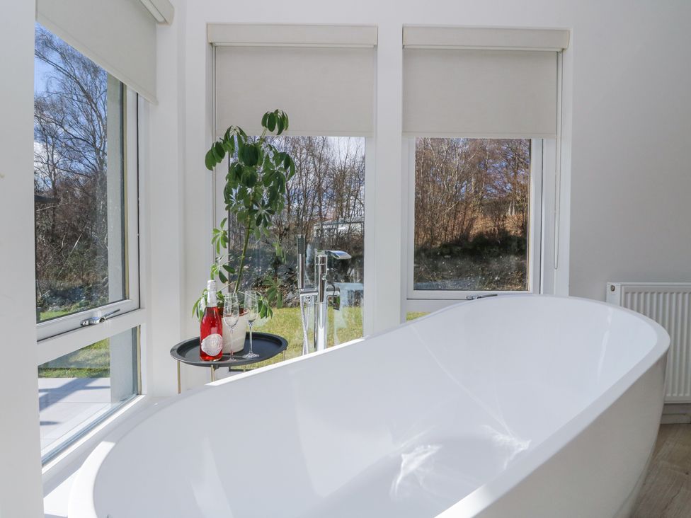 A white bathtub near windows with roller blinds a small round table with a plant bottle and glasses at Macgregor Ranch in Achnabobane near Spean Bridge
