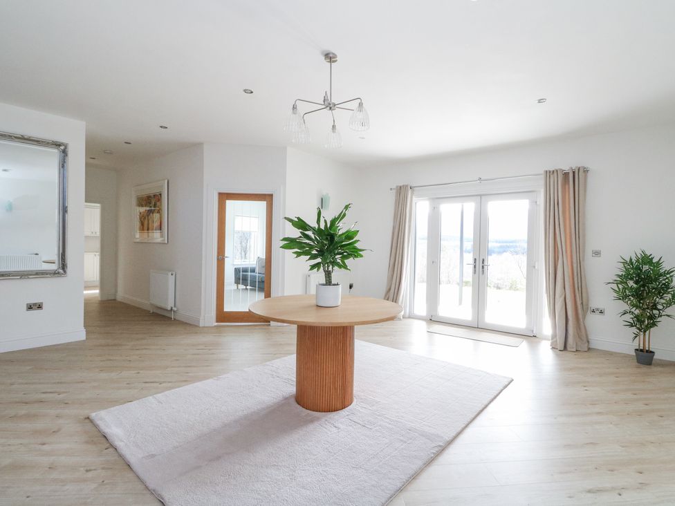 A room with a round wooden table on a rug with a plant on top and glass doors with curtains at Macgregor Ranch in Achnabobane near Spean Bridge