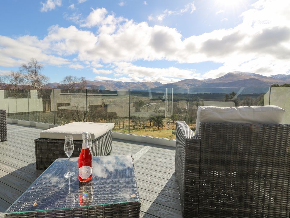 An outdoor patio with wicker furniture a glass table with a bottle and two glasses and mountains in the background at Macgregor Ranch in Achnabobane near Spean Bridge