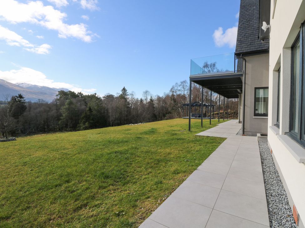 A tiled patio beside a modern house with a glass balcony and grassy field with trees and mountains in the background at Macgregor Ranch in Achnabobane near Spean Bridge