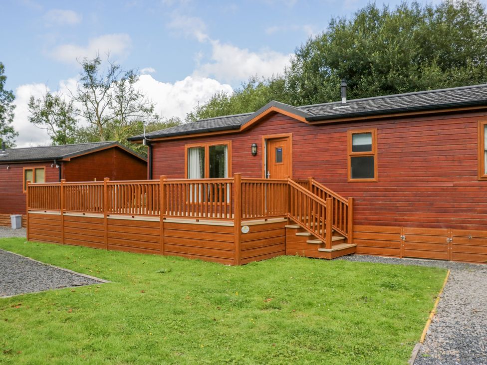 A wooden lodge with a deck and stairs in front of a grassy area at Waterside Lodge 55A Bowness White Cross Bay near Windermere