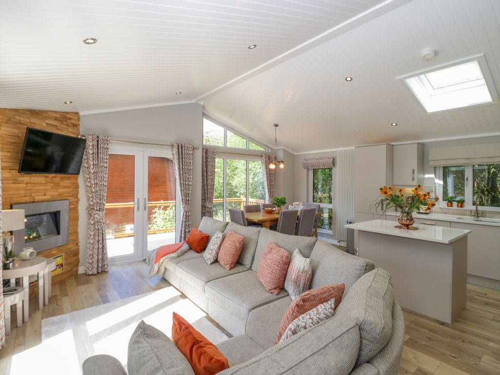 An open living area with a grey sofa and patterned cushions a dining table with chairs and a kitchen island with flowers at Waterside Lodge 55A Bowness White Cross Bay near Windermere