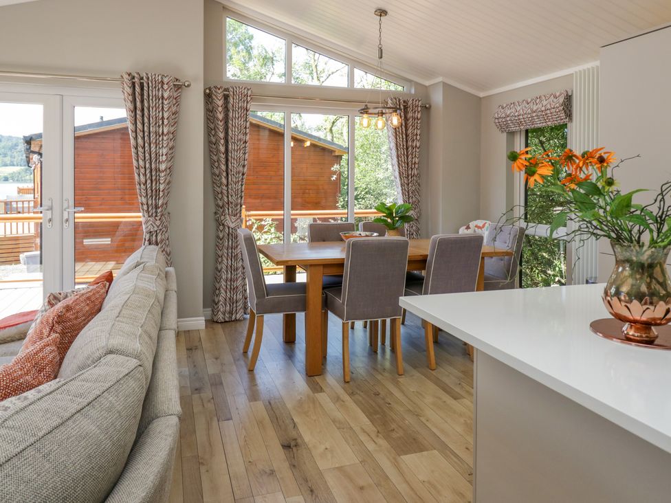 A dining area with six chairs around a wooden table next to large windows and a sofa in a living space at Waterside Lodge 55A Bowness White Cross Bay near Windermere