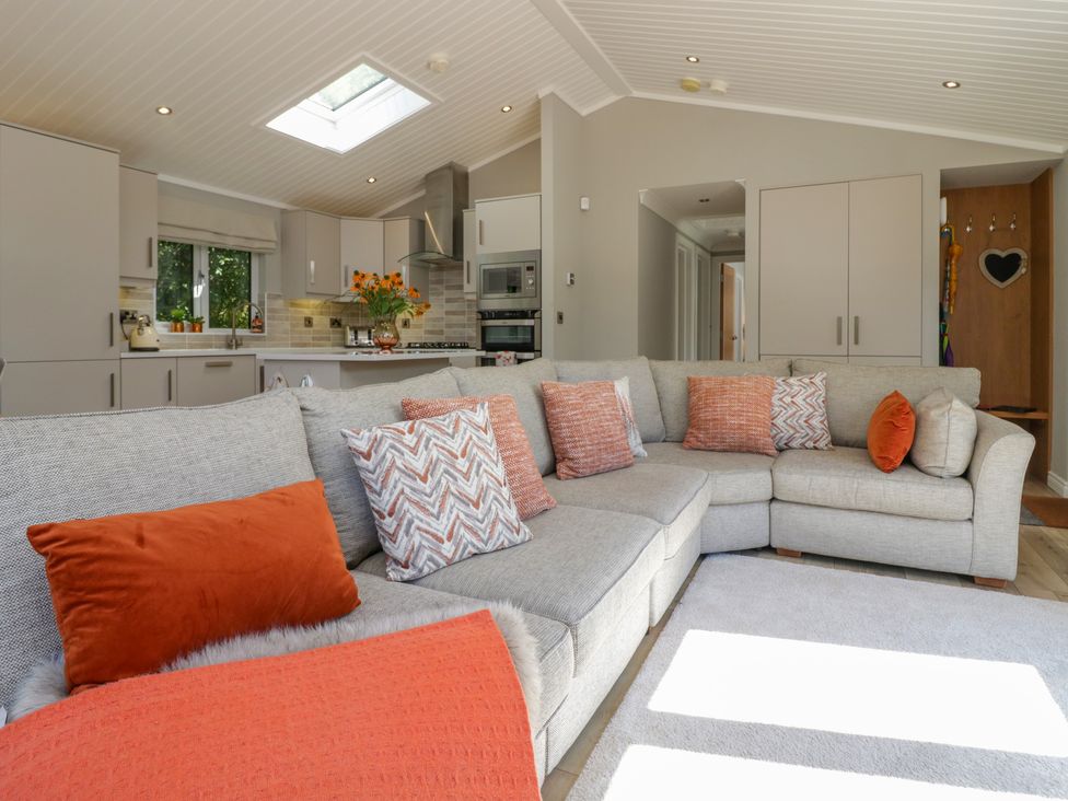 A living room with a large gray sectional sofa with orange and patterned cushions and a kitchen with gray cabinets and appliances at Waterside Lodge 55A Bowness White Cross Bay near Windermere