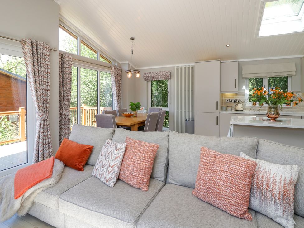 A living room with a grey sofa with cushions and blanket next to a dining table with chairs and a kitchen counter with flowers at Waterside Lodge 55A Bowness White Cross Bay near Windermere