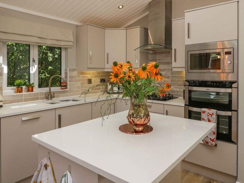 A kitchen with a white island countertop holding a vase of orange flowers and beige cabinets with built-in stainless steel oven and microwave at Waterside Lodge 55A Bowness White Cross Bay near Windermere
