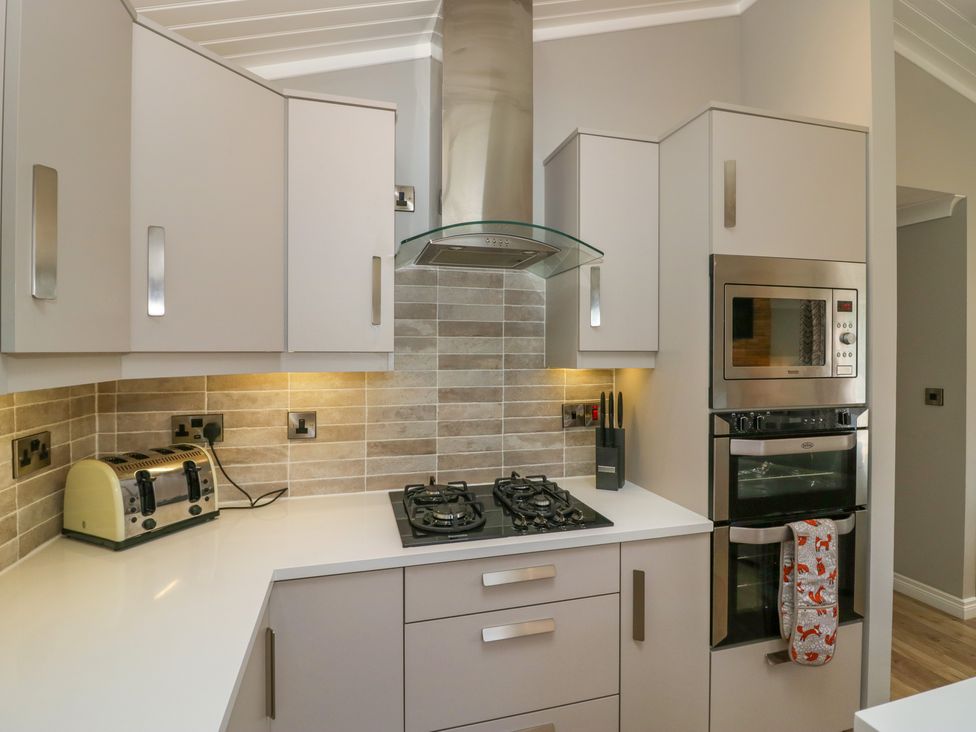 A kitchen with white cabinets a toaster a gas stove a range hood a knife block a built in microwave and oven and an oven mitt at Waterside Lodge 55A Bowness White Cross Bay near Windermere