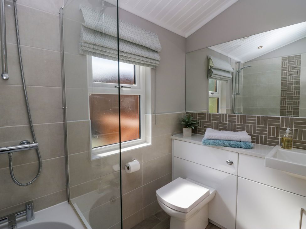 A bathroom with a shower over a bathtub a window with a blind a toilet and a sink with towels and a plant on the counter at Waterside Lodge 55A Bowness White Cross Bay near Windermere