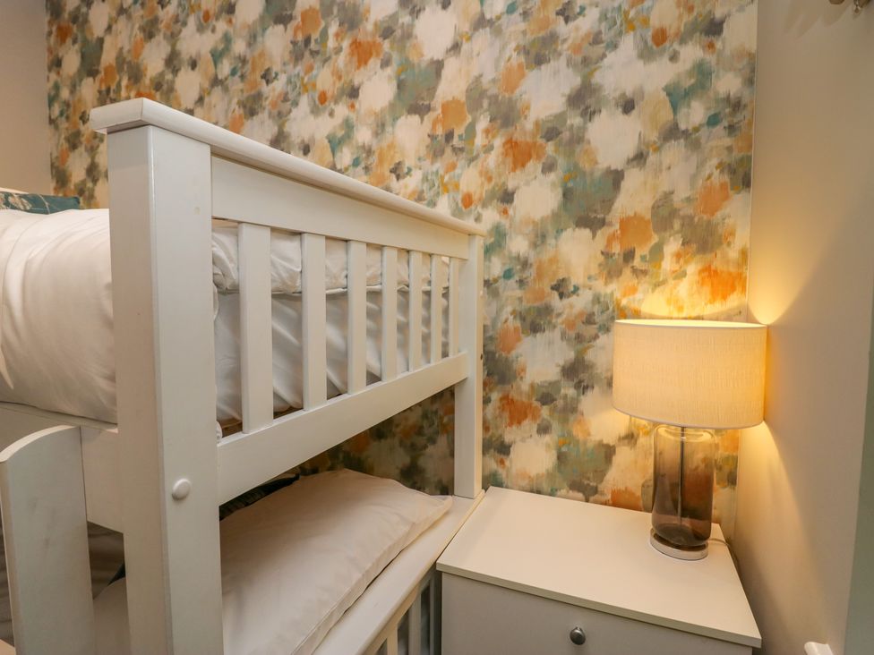 A white bunk bed next to a white nightstand with a lamp and a multicolored abstract wallpaper in a bedroom at Waterside Lodge 55A Bowness White Cross Bay near Windermere