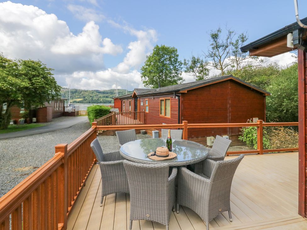 An outdoor deck with a round glass table and six wicker chairs near wooden buildings at Waterside Lodge 55A Bowness White Cross Bay near Windermere