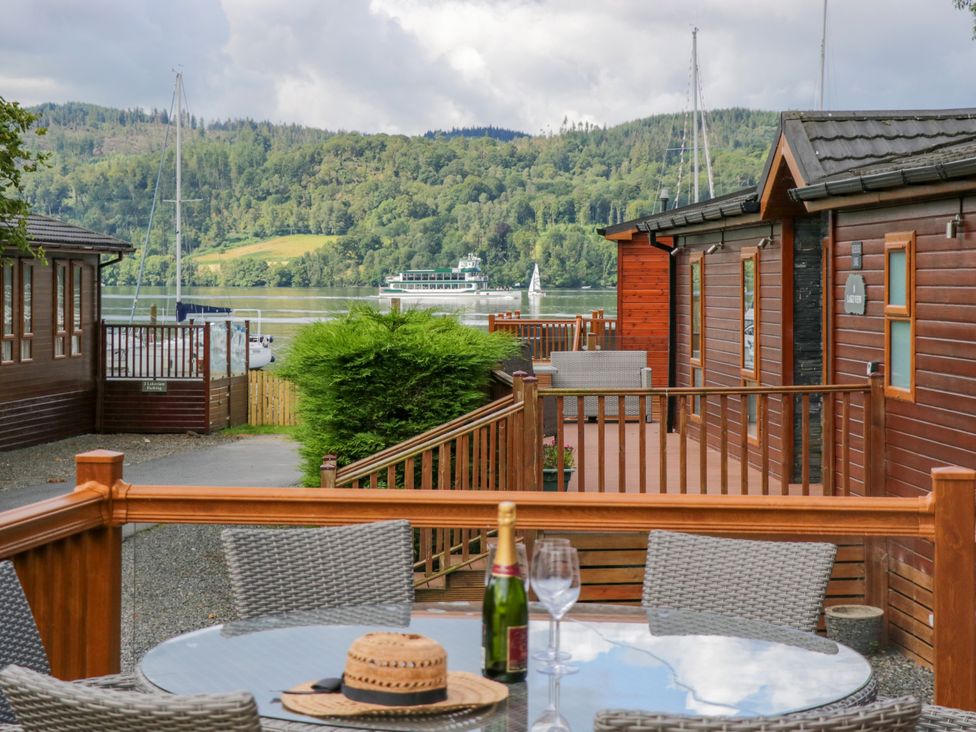 A waterside patio with a round glass table set with a bottle of champagne and hat between wooden lodges at Waterside Lodge 55A Bowness White Cross Bay near Windermere