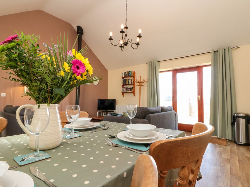 A dining table with a polka dot tablecloth and flowers in a vase in a room with a gray sofa a TV wooden chairs curtains and a chandelier at Partridge Cottage neaer Filey