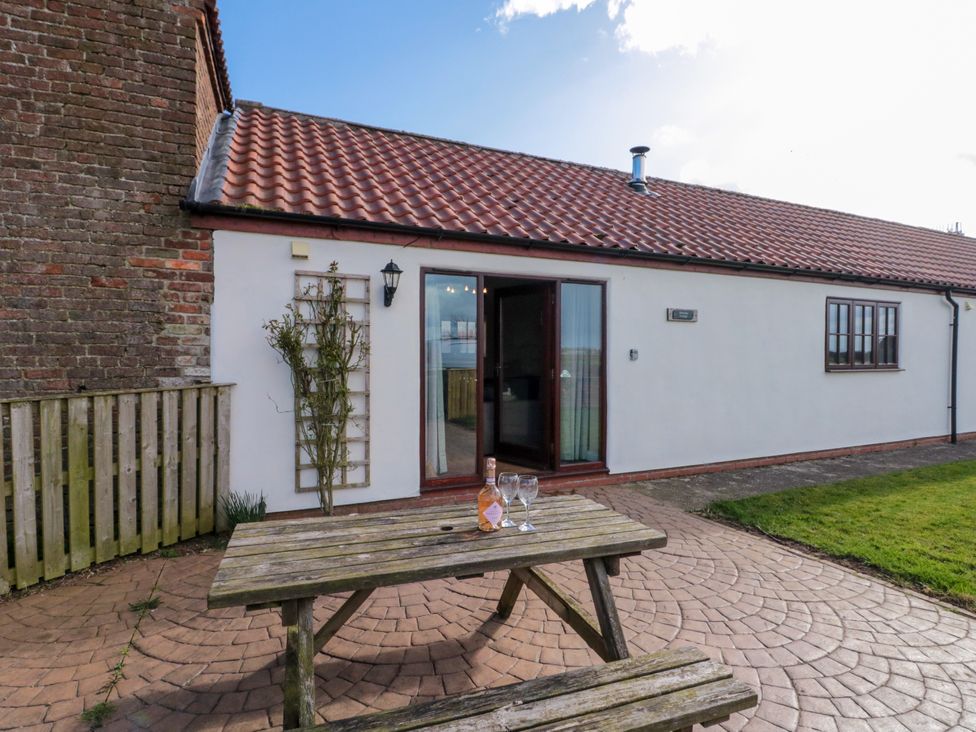 A wooden picnic table with a bottle and two glasses on a paved patio outside a white single-story building with a tiled roof and glass door at Partridge Cottage neaer Filey
