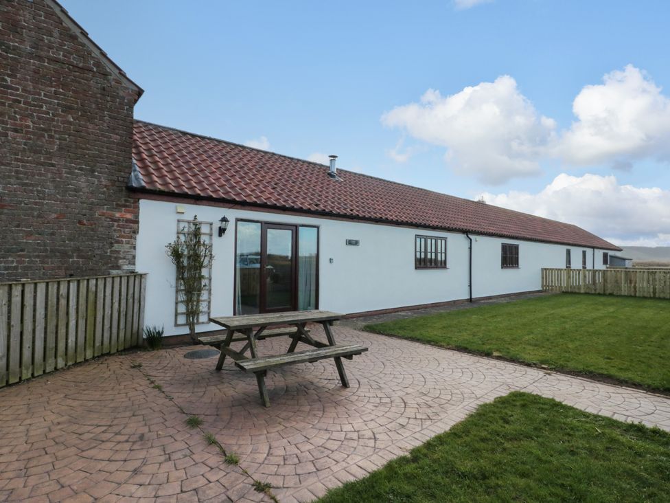 An outdoor patio area with a wooden picnic table and a grassy lawn next to a white building with tiled roof at Partridge Cottage neaer Filey