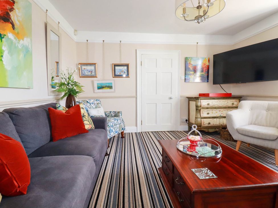 A living room with a gray sofa red cushions striped carpet wooden coffee table armchairs paintings on the walls and a TV at Cathedral View Cottage in Truro