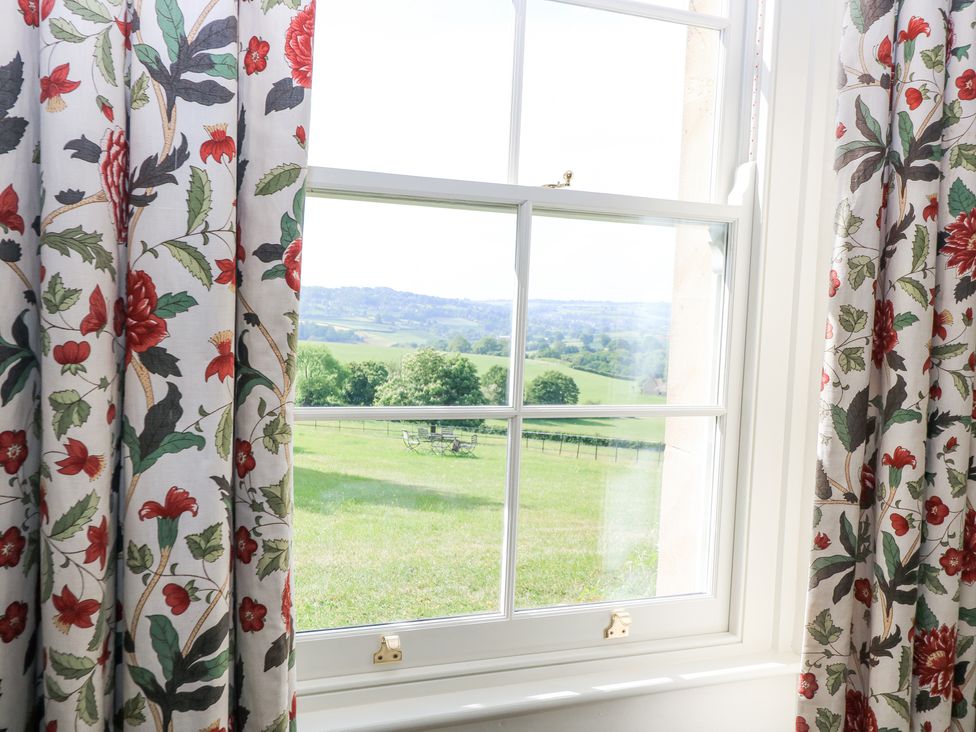 A window with floral curtains looking out to a green field with distant hills at Peartree in Colerne
