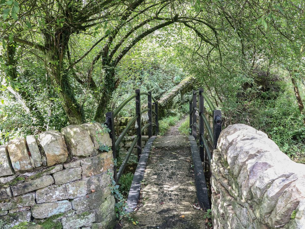 A bridge crossing a pathway with trees and stone walls at Brook Cottage in Hayfield, High Peak