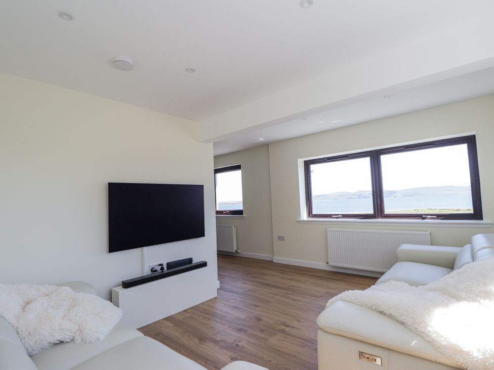 A living room with two white sofas a wall-mounted TV and large windows overlooking water at Calmdale Inverasdale near Poolewe
