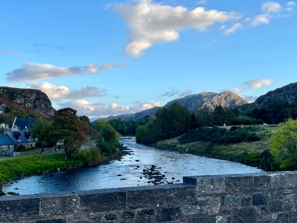 A river flowing between green trees and hills with clouds in the sky and stone buildings on the left side