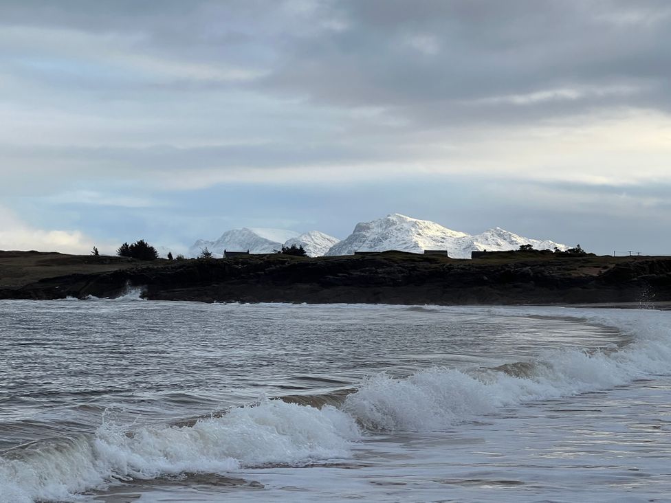 Waves breaking on a shoreline with rocky cliffs and snow-covered mountains in the background at Calmdale in Inverasdale near Poolewe