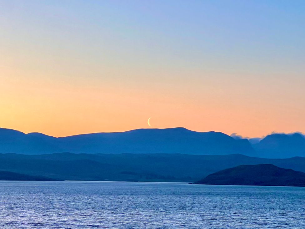 A crescent moon above mountains with a body of water in the foreground during sunset at Calmdale Inverasdale near Poolewe