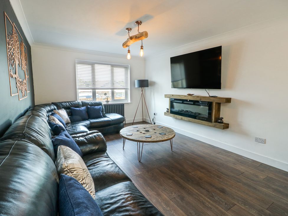 A living room with a black leather corner sofa a round wooden coffee table a wall-mounted TV and an electric fireplace at Ty Copr in Amlwch