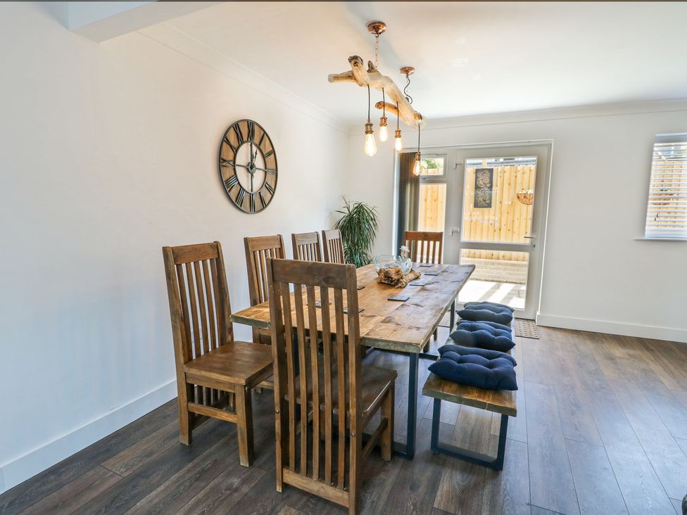 A dining room with a wooden table surrounded by six chairs and a bench with blue cushions a large wall clock and a glass door at Ty Copr in Amlwch