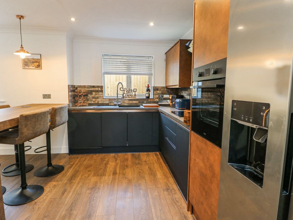 A kitchen with a wooden floor, black lower cabinets, wooden upper cabinets, a built-in oven, refrigerator, sink under a window, and a breakfast bar with stools at Ty Copr in Amlwch