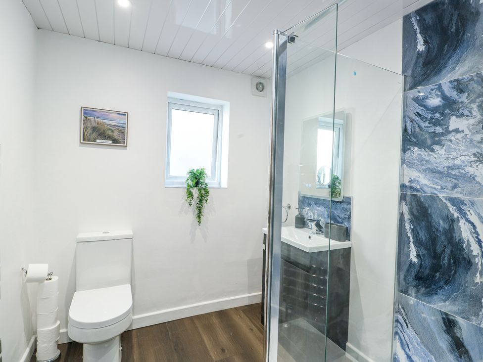A bathroom with a toilet, a small window, a plant on the window ledge, a framed picture on the wall, a sink with a mirror, and a shower with blue marbled tiles at Ty Copr in Amlwch