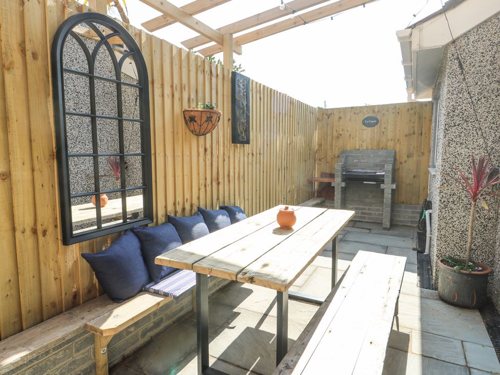An outdoor patio area with wooden benches and table along a wooden fence with cushions and a wall mirror and a barbecue grill and potted plants at Ty Copr in Amlwch